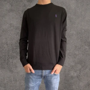 Svart slim fit tröja Polo Ralph Lauren - Svart långärmad tröja från Polo Ralph Lauren i slim fit med stretch. Klassisk rund halsringning och lila broderad logga på bröstet. Perfekt för dig som gillar stilren och enkel design med en touch av färg.