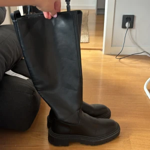 Svarta höga boots - Säljer ett par svarta höga boots med rund tå och platt sula. Dom har en chunky, robust look som passar perfekt till jeans eller kjol. Stövlarna är stilrena och har en minimalistisk design utan synliga detaljer eller mönster.