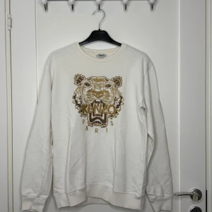 Kenzo Crewneck – Krämfärgad (M) - Stilren crewneck från Kenzo i en fräsch krämfärg. Mjuk och bekväm passform med subtila detaljer – lätt att styla både uppklätt och avslappnat. Passar storlek M, normal till lite mindre i fit.  👕 Klassisk modell med rund hals 🎨 Krämfärgad – passar allt 🧵 Skön kvalitet och snygg passform