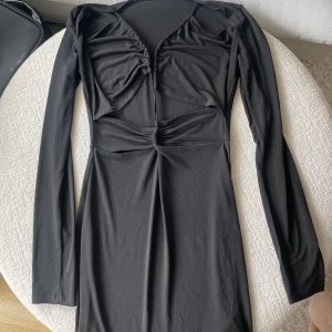 Svart långärmad klänning med cut-outs - Snygg svart kortklänning med långa ärmar och coola cut-outs framtill. Klänningen har rynkade detaljer vid bysten och midjan som ger en trendig look. Perfekt för dig som gillar en stilren men edgy outfit.