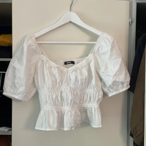 Vit blus med puffärm från BikBok - Supersöt vit blus från BikBok med puffiga korta ärmar och smockad midja. Blusen har en söt spetskant vid urringningen och volang nertill. Perfekt för en romantisk look och passar till både jeans och kjol.