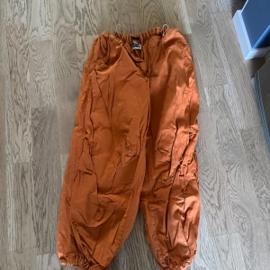 Orange cargopants från Reclaimed Vintage - Säljer ett par orange cargopants från Reclaimed Vintage. Byxorna har loose fit, resår i midjan och benslut samt flera fickor på sidorna. Perfekta för en avslappnad och trendig streetstyle-look.