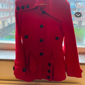 Röd kappa med stora knappar, stl 36 - Snygg röd kappa i klassisk modell med stora svarta knappar och bred krage. Jackan har långa ärmar, fickor med lock och markerad midja. Perfekt för dig som vill sticka ut med färg i höst och vinter. Passar dig som gillar en stilren men ändå cool look.