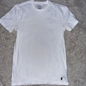 Vit t-shirt från Polo Ralph Lauren - Klassisk vit t-shirt från Polo Ralph Lauren med rund hals och korta ärmar. Tillverkad i mjuk bomull och har den ikoniska lilla broderade loggan i mörkblått vid nederkanten. Perfekt basic-plagg med clean look.