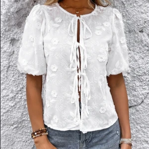 Vit blommig blus med knytband - Superfin vit blus med broderade blommor och puffiga korta ärmar. Blusen har flera knytband framtill och är lätt transparent med romantisk känsla. Perfekt att styla med jeans eller kjol för en trendig look.