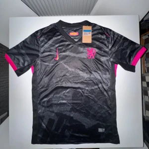  Nike x Chelsea fotbollströja  - Chelsea fotbollströja från Nike i svart med subtilt mönster och rosa detaljer. Broderad Nike-logga och Chelsea-lejon på bröstet. Kortärmad modell i lätt, andningsbart Dri-FIT-material. Perfekt för dig som vill sticka ut på planen eller läktaren.