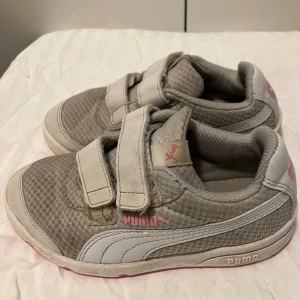Grå och rosa sneakers från Puma - Snygga sneakers från Puma i grått med vita och rosa detaljer. Skorna har två praktiska kardborreband, mesh-yttermaterial och klassisk Puma-logga på sidan och tungan. Använda men fullt fungerande ett tag till.