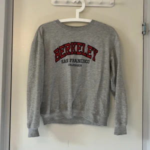 Grå college-tröja Berkeley 157 - Grå sweatshirt från 157 med tryck 'Berkeley San Francisco California' i rött och svart framtill. Klassisk rund hals, ribbade muddar och lång ärm. Perfekt för en chill och avslappnad stil. Lite sliten på ena ärmen (se bild 4) men annars i fint skick.