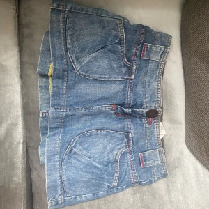 Jeanskjol  - Snygg kort jeanskjol med coola röda sömmar och stora fickor fram. Kjolen har breda hällor för bälte och klassisk knappstängning. Perfekt för dig som gillar en avslappnad men trendig stil.