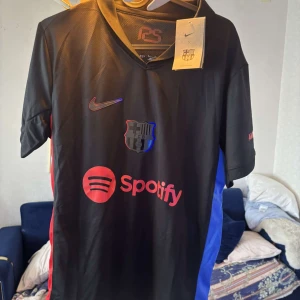 Svart FC Barcelona Nike fotbollströja - Snygg svart FC Barcelona fotbollströja från Nike med klubbmärke och Spotify-logga på bröstet. Tröjan har röda och blå sidopaneler, korta ärmar och V-ringning. Materialet är lätt och andas, perfekt för träning eller match.