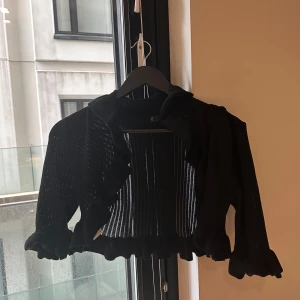 Svart bolero från mango - Säljer en svart, ribbad kofta/bolero från Mango med volangkant längs ärmslut och nederkant. Koftan är croppad och har långärmade ärmar. Perfekt att slänga över en topp för en snygg layering-effekt.