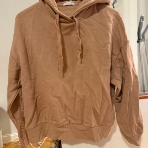 Beige hoodie från NewYorker  - Säljer en beige hoodie från NewYorker i storlek S med snörning i halsen och stor huva med tryck baktill. Tröjan har en relaxed passform och är gjord i mjuk bomullsmix.