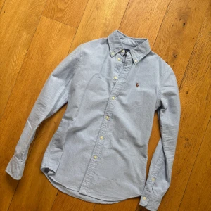 Ralph Lauren Skjorta (Pris diskuterbart) -  Säljer en klassisk ljusblå skjorta i ny skick från Ralph Lauren i SLIMFIT(står custom fit men sitter som slim)  med button-down krage och broderad logga på bröstet. Storlek S fast liten i Storlek .Skjortan har lång ärm, normal passform och är tillverkad i mjuk bomull. Perfekt till jeans eller chinos för en clean look. !! PRIS ÖR DISKUTERBART !!