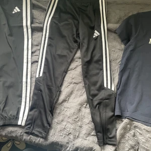 Svarta Adidas mjukisbyxor med ränder - Svarta mjukisbyxor från Adidas med klassiska vita ränder längs sidorna och Adidas-logga på låret. Byxorna har elastisk midja med snörning och är tillverkade i ett mjukt syntetmaterial. Perfekta för träning eller chill.