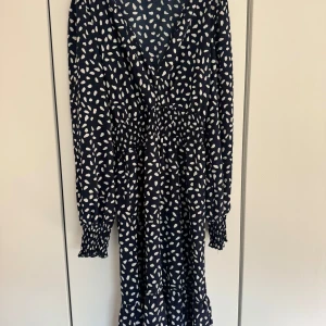 Mörkblå kortklänning med vita prickar  - Snygg kortklänning  i mörkblått med vita prickiga mönster. Klänningen har v-ringning, långa ärmar med smock vid handlederna och markerad midja med resår. Använd 1-2 gånger, är som ny!! Priset går att diskutera 