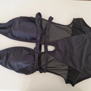 Svart baddräkt med mesh från NA-KD - Snygg svart swimsuit från NA-KD med trendiga meshdetaljer och coola utskärningar. Överdelen har trekantskupor och knyt i nacken, medan underdelen har smala band och genomskinliga partier. Perfekt för dig som vill sticka ut på stranden! 