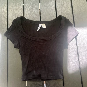 Svart croppad topp från H&M Divided - Svart ribbad croppad topp från H&M Divided med korta ärmar och rundad, djup urringning. Perfekt att matcha med höga jeans eller kjol för en chill och trendig look.