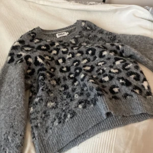Grå leopardmönstrad stickad tröja ONLY - Supermysig grå stickad tröja från ONLY med leopardmönster i svart och vitt. Tröjan har lång ärm, ribbade muddar och en loose fit som ger en chill vibe. Perfekt för kalla dagar när du vill vara både snygg och bekväm.