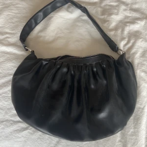 Svart axelväska från Gina Tricot - Snygg svart axelväska från Gina Tricot i mjukt skinnimitation. Väskan har en rundad form med rynkade detaljer och bred axelrem. Stängs med dragkedja och har ett innerfack med extra dragkedja. Perfekt att ha till skolan då den rymmer det mesta. Bara att höra av dig om du har frågor! 