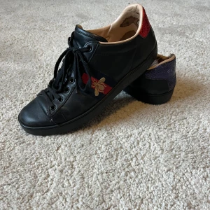 Gucci sneakers med broderad bi - Svarta sneakers från Gucci i skinn med klassiska röd-blå ränder och broderad guldfärgad bi på sidan. Baksidan har en röd och en lila häl i ormskinnsmönster med Gucci-logga. Skorna har snörning och platt sula, perfekt för dig som vill sticka ut.