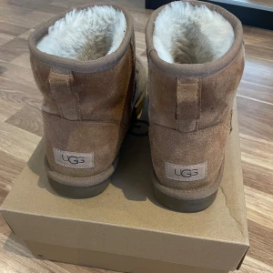 Bruna UGG boots i mocka med fårfoder - Mysiga bruna UGG boots i mocka med mjukt vitt fårfoder på insidan. Klassisk rund tå och platt sula, perfekta för kalla dagar. Ikonisk UGG-logga baktill och enkel slip-on-modell. Passar dig som vill ha både stil och komfort under vintern.