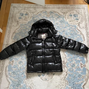 Svart Moncler pufferjacka med huva - Snygg svart pufferjacka från Moncler med glansig finish och huva. Jackan har dragkedja framtill, två fickor med dragkedja och klassisk Moncler-logga på ärmen. Perfekt för kalla dagar när du vill hålla stilen och värmen.