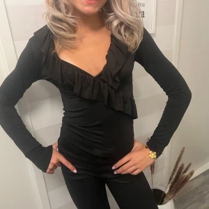 Svart långärmad topp med volang - Snygg svart långärmad topp med djup v-ringning och volangdetalj framtill. Figurnära passform som framhäver siluetten. Perfekt för dig som gillar stilrena plagg med en twist. Mjuk och stretchig känsla.