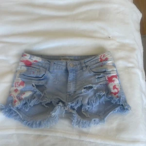 Ljusblå slitna jeansshorts Mango - Supercoola ljusblå jeansshorts från Mango med råa, fransiga kanter och slitningar. Shortsen har röda och vita färgstänk på fram- och baksidan för en edgy vibe. Klassisk femficksmodell och låg midja. Perfekta för sommaren och festivaler! 