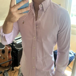 Ljusrosa skjorta från Ralph Lauren - Säljer en klassisk ljusrosa skjorta från Ralph Lauren med button-down krage och broderad logga på bröstet. Skjortan har lång ärm, normal passform och är tillverkad i mjuk bomull. Perfekt för dig som gillar stilrena och tidlösa plagg.
