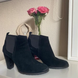 Svarta mocka chelsea boots med klack - Snygga svarta chelsea boots i mocka med bred resår på sidorna och rejäl hög klack. Skorna har rund tå och en klassisk siluett som passar till många outfits. Perfekta för dig som vill ha en stilren och trendig look. Storlek 40