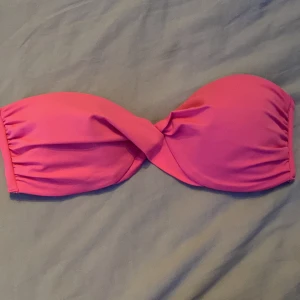 Rosa bandeau bikiniöverdel - Snygg bandeau bikiniöverdel i stark rosa färg med twistad front. Axelbandslös modell med vadderade kupor och smidig metallknäppning bak. Perfekt för dig som vill ha en enkel men trendig look på stranden.