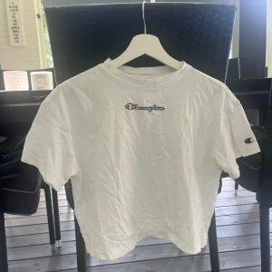 Vit Champion t-shirt med broderad logga - Vit t-shirt från Champion med broderad logga på bröstet och ett mindre Champion-märke på ärmen. Klassisk passform och rund halsringning. Perfekt basic-plagg för en clean och sportig stil.