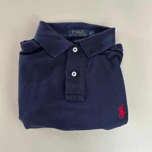 Marinblå pikétröja från Ralph Lauren - Klassisk marinblå pikétröja från Ralph Lauren i bomull. Tröjan har korta ärmar, krage med två knappar och den ikoniska röda polospelaren broderad på bröstet. Custom fit-modell som sitter snyggt och passar perfekt till jeans eller shorts.