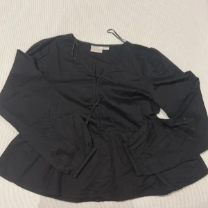 Svart blus med rosetter och volang  - En svart blus med volanger och v-ringad krage, rosetter på framsidan. Storlek 158-164, sms för mer bilder och bilder med tröjan på och priset kan diskuteras och sänkas.