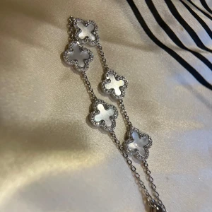 Silverfärgat armband med fyrklöver - Superfint armband i silverfärg med flera fyrklöverformade länkar. Varje klöver har en skimrande vit pärlemoryta och är kantad av små glittrande stenar. Kedjan är tunn och elegant, perfekt för att ge en lyxig touch till din stil. Around 21cm