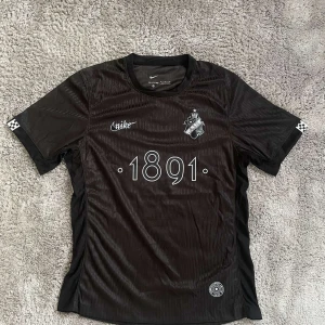 Svart AIK fotbollströja Nike 1891 - AIK fotbollströja i svart från Nike med trycket '1891' på bröstet och klubbmärket på vänster sida. Tröjan har korta ärmar, diskreta ränder i tyget och små rutiga detaljer vid ärmsluten. Materialet är lätt och andas, perfekt för match eller träning.