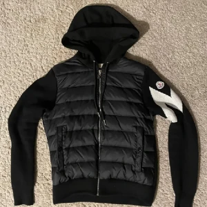 Moncler Cardigan - Snygg svart pufferjacka från Moncler med huva och dragkedja. Jackan har quiltad front i glansigt material, stickade ärmar och rygg samt vit detalj på ena ärmen. Perfekt för dig som vill ha en sportig och trendig look.