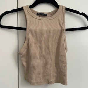 Beige ribbad linnetopp från Zara - Snygg beige croppad linnetopp från Zara med ribbad struktur. Toppen är ärmlös och har en klassisk rund halsringning. Perfekt att matcha med jeans eller kjol för en clean och trendig look.
