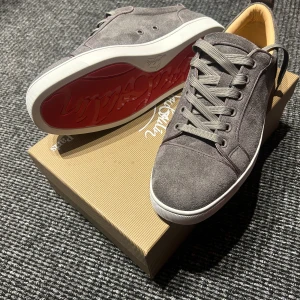 Gråa Louboutin skor  - Snygga grå sneakers i mocka från Christian Louboutin med klassisk röd sula. Skorna har rund tå, snörning och vita kanter på sulan. Insidan är beige och de har en stilren, minimalistisk design. Priset kan diskuteras 😁