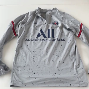 PSG x Jordan långärmad fotbollströja - Snygg långärmad PSG fotbollströja från Jordan i ljusgrått med mörkblå stänk, broderad PSG-logga och Jordan Jumpman på bröstet. Tröjan har dragkedja vid halsen och röda detaljer på ärmarna. Tillverkad i lätt och ventilerande funktionsmaterial.