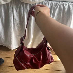 Röd axelväska dragkedjor - Cool röd axelväska i skinn med bred axelrem och silvriga detaljer. Väskan har två dragkedjor framtill och en snygg spänndetalj på remmen. Perfekt storlek för det viktigaste . Köpt på behrska i Rom.