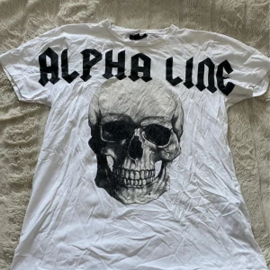 Alpha Line vit t-shirt Machine World Tour - Vit t-shirt från Alpha Line med stor svart text framtill och en cool dödskallegrafik. På ryggen finns turnédatum och städer tryckta i svart, samt texten 'Machine World Tour 2021'. Klassisk passform och rund hals.