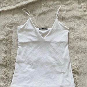 Vit linnetopp från Zara - Säljer en stilren vit linnetopp från Zara med smala axelband och v-ringning. Toppen är i ett mjukt, slätt material som känns lyxigt och passar perfekt till jeans eller kjol. Enkel och clean design som är lätt att matcha.