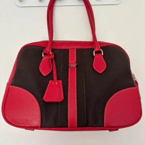 Prada Bowling bag - AuthenticPrada Handbag – Red Leather & Brown Canvas  Size: 25 × 31 × 12 cm  Condition: almost as new, no visible signs of wear  A striking and rare Prada Bauletto (bowling-style) handbag in a bold red and chocolate colorway. Crafted with structured precision, this piece pairs timeless materials with sporty elegance: • Material: Sturdy brown canvas body with contrasting red textured leather trim and handles • Hardware: Silver-tone details, including a front buckle accent and top zip closure • Log