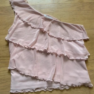 Ljusrosa volang topp oneshoulder - Supersöt ljusrosa topp med flera volanger och oneshoulder-design. Tunt och lätt material som ger en luftig känsla. 
