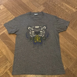Grå Kenzo t-shirt med tigertryck - Snygg grå t-shirt från Kenzo med ikoniskt tigertryck och Kenzo Paris-logga på bröstet. Klassisk rund hals och korta ärmar. Perfekt för dig som vill sticka ut med en streetstyle-vibe. Materialet känns mjukt och skönt mot huden. Storlek Small, i mycket bra skick. 