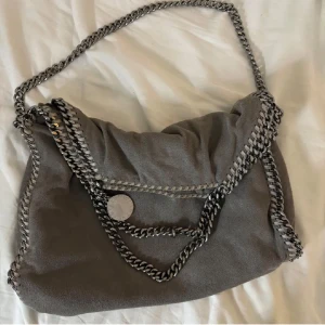 Stella McCartney väska - Säljer nu min fina gråa axelväska från Stella McCartney Falabella i mellan storlek. Väskan har silvriga kedjdetaljer och en rimlig insida med rosa foder. Den är i fint skick och använd endast ett fåtal gånger! Ordinarie pris är ca 12.000 kr, en av sidorna har ett minimalt hål som knappt syns vid användning (se sista bild), därav priset. Vid fler bilder eller äkthetsbevis är det bara att kontakta! 