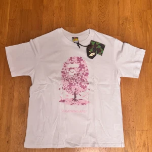 Vit BAPE t-shirt med rosa blomtryck - Vit t-shirt från A Bathing Ape i storlek L med ikoniskt BAPE-ansikte i rosa och lila blommor på bröstet. Klassisk passform och korta ärmar. Tillverkad i mjuk bomull och har originaltaggar kvar. Perfekt för dig som gillar streetwear och unika prints.