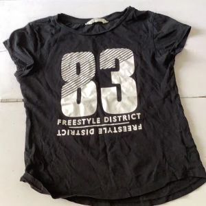 Svart t-shirt med tryck från H&M - Svart t-shirt från H&M med stort vitt tryck '83 Freestyle District' på framsidan. Modellen är klassisk med rund hals och korta ärmar. Perfekt för en avslappnad och sportig stil.