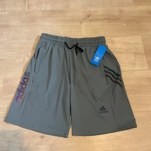 Gröna träningsshorts från Adidas Originals - Snygga gröna shorts från Adidas Originals med svart snörning i midjan och klassiska Adidas-loggan på ena benet. Färgade ränder längs sidan och tryckt Adidas-text. Perfekta för träning eller en sportig stil.